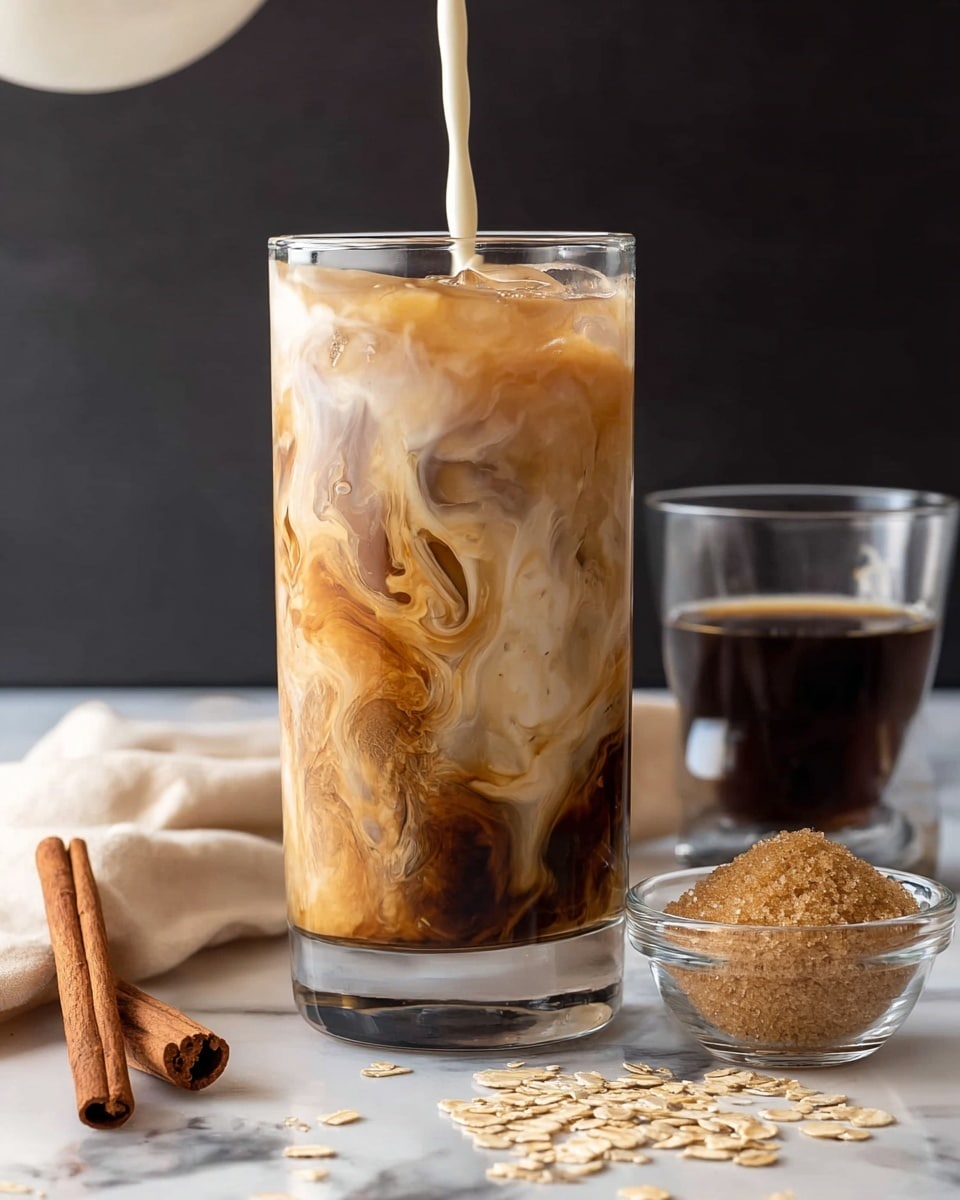 A tall clear glass filled with iced coffee shows three layers: the bottom layer is light creamy beige, the middle layer is a soft brown with swirls mixing with the cream, and the top layer is frothy light brown foam with visible ice cubes floating. Two light wooden straws stand inside the glass. The glass rests on a white marble coaster on a dark wooden table. Around the glass are cinnamon sticks, scattered oat grains, a small clear glass cup of dark espresso, and another small glass bowl filled with light brown sugar. The background is a soft beige cloth against a dark blurry wall. photo taken with an iphone --ar 4:5 --v 7
