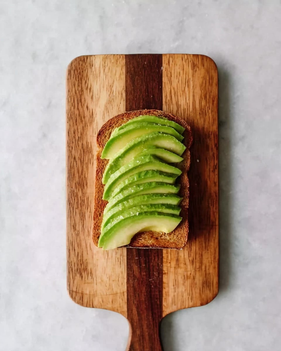 A wooden cutting board with three different shades of brown holds a single slice of toasted bread topped with six evenly arranged, bright green avocado slices. The foam-like white marbled surface beneath the board contrasts with the warm tones of the toast and cutting board, creating a clean and fresh look. photo taken with an iphone --ar 4:5 --v 7