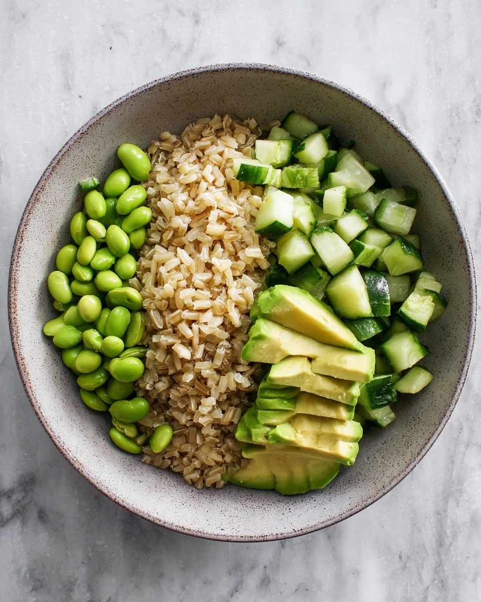 A bowl with a base layer of brown rice showing grains with a soft texture and light brown color, topped with three distinct sections: one section of bright green edamame beans with a smooth, slightly shiny surface, another section of diced cucumber pieces showing dark green skin and light green inside, and the last section of neatly sliced avocado with soft, light green flesh and darker green edges, all arranged side by side in the bowl, placed on a white marbled surface. Photo taken with an iphone --ar 4:5 --v 7
