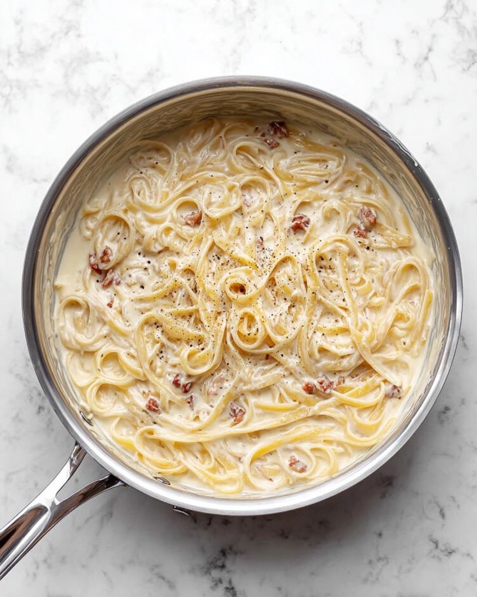 The image shows a silver pan filled with creamy pasta. There are many long, flat noodles coated in a thick, white sauce with small pieces of browned bits scattered throughout. The sauce looks smooth and slightly shiny, with a little black pepper sprinkled on top. The pan sits on a white marbled surface that adds a clean and simple background to the dish. photo taken with an iphone --ar 4:5 --v 7