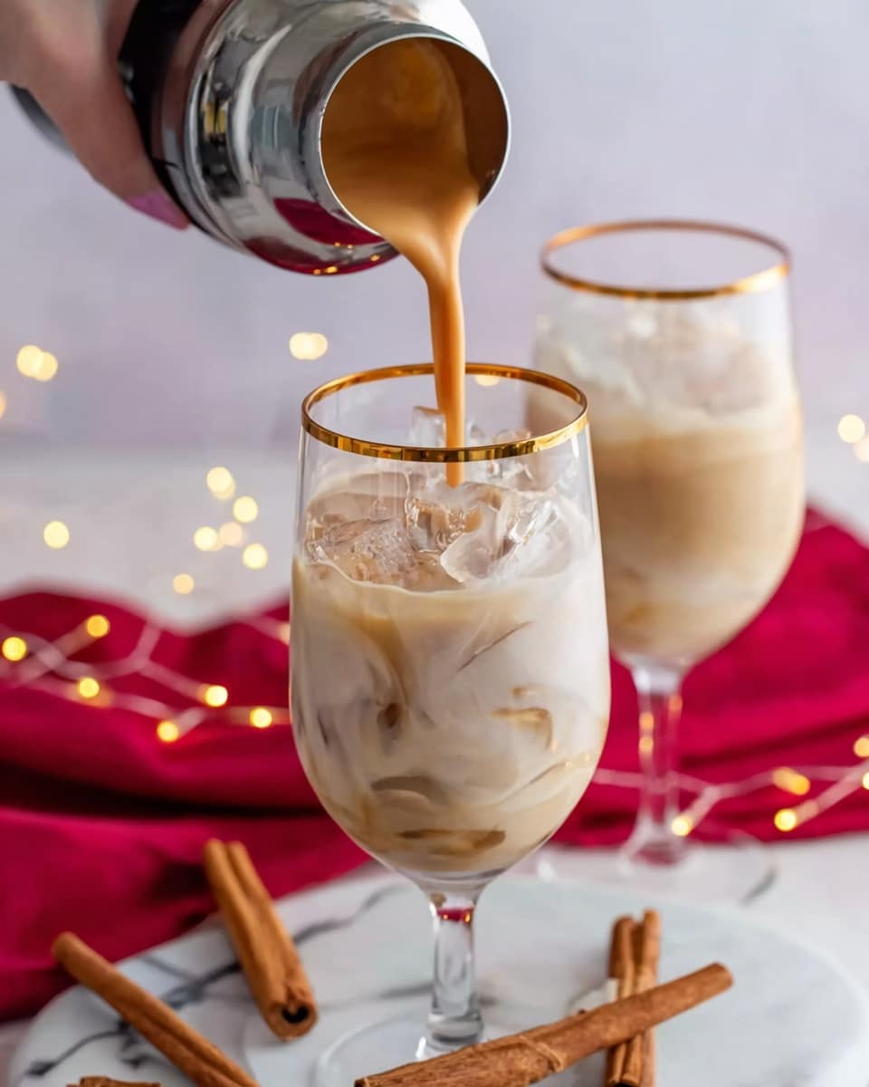 The image shows two tall white glasses with golden rims, filled with a creamy light brown drink and ice cubes. One glass is in the foreground, being filled from a shaker held by a woman's hand at the top left. The creamy drink is pouring smoothly into the glass, creating a soft swirl on the surface. In the background, there is another similar glass also containing the drink. The glasses are placed on a white marbled surface. Around them, there are several brown cinnamon sticks and a red cloth spread casually. Small warm fairy lights add a cozy feel, with soft bokeh in the background. photo taken with an iphone --ar 4:5 --v 7
