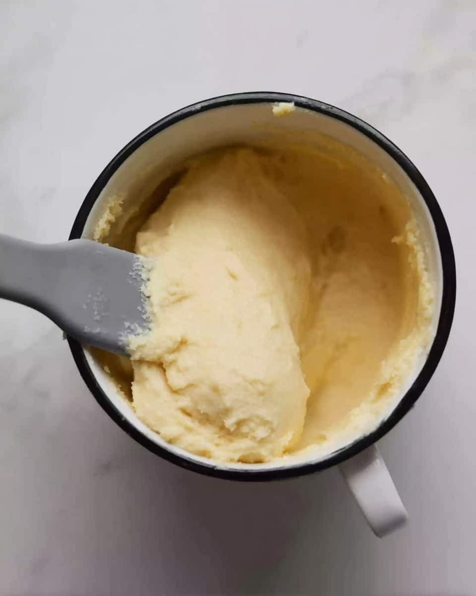 A white cup with a dark rim contains a light yellow, smooth dough being pressed by a gray spatula. The dough forms one thick, soft layer resting against the side of the cup, with some dough stuck to the spatula. The background is a white marbled texture. Photo taken with an iphone --ar 4:5 --v 7