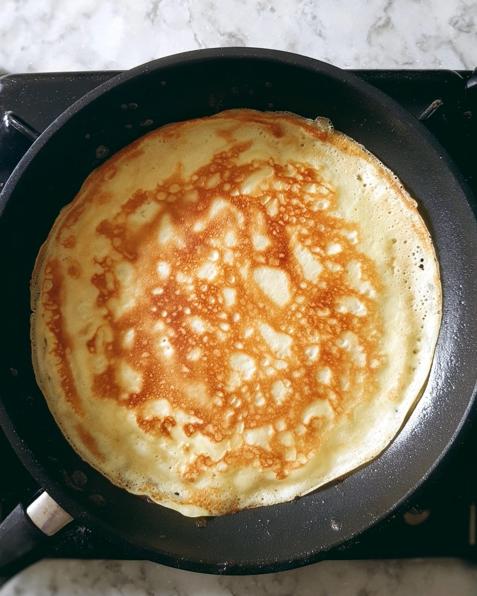 A single light golden crepe is cooking in a black frying pan on a stove, showing slight browning spots with a bubbly texture across its surface. The crepe has a round shape with thin edges that are slightly crisp. The pan sits on black stove grates, and some light reflections can be seen. The background surface is a white marbled texture. photo taken with an iphone --ar 4:5 --v 7