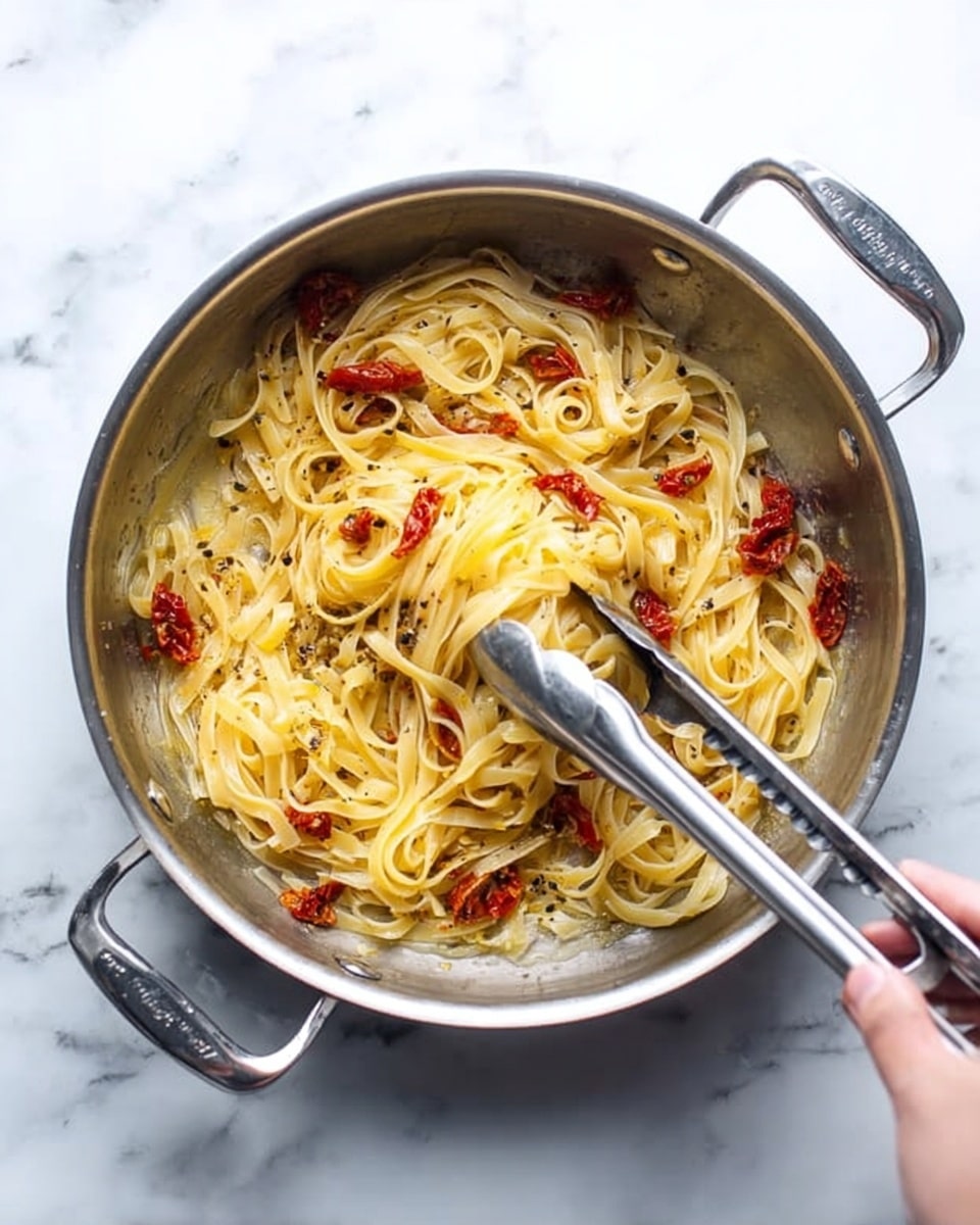 A metal pan filled with yellow flat noodles mixed with small pieces of red sun-dried tomatoes, sitting on a white marbled surface. The noodles are spread unevenly in the pan, showing some oily liquid with black pepper specks. A woman's hand holds silver tongs, lifting some noodles from the pan. The pan has two silver handles on each side. photo taken with an iphone --ar 4:5 --v 7