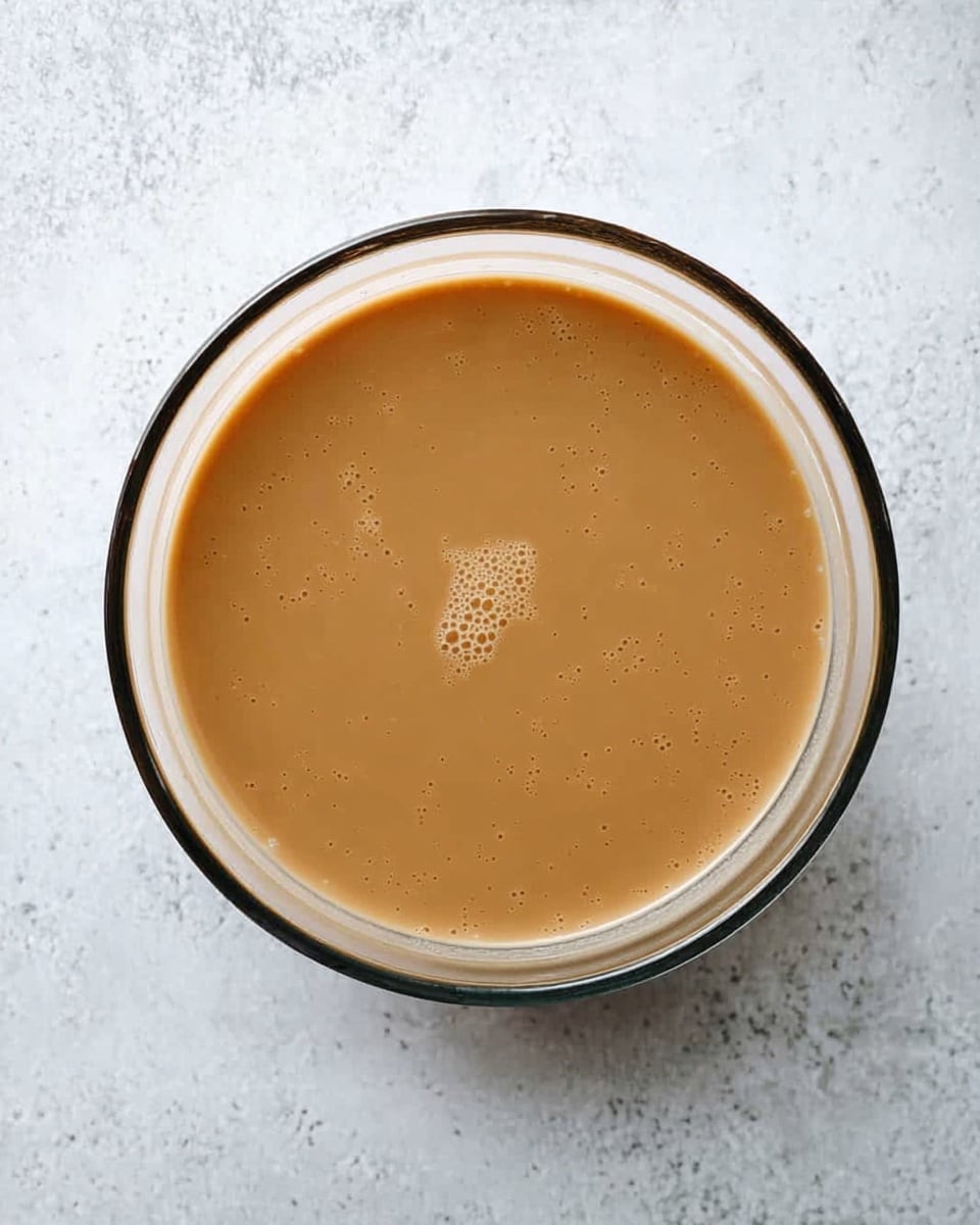 A clear glass bowl is filled with a smooth, light brown sauce or batter, resting in the center on a white marbled surface. The liquid looks creamy with a few small bubbles spread near the middle, showing a soft texture. The bowl has a thin black rim around the top, making the light brown mixture inside stand out clearly. photo taken with an iphone --ar 4:5 --v 7