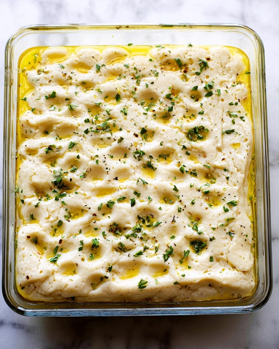 The image shows a clear rectangular glass dish filled with a dough-based dish that has a thick, pale golden top layer with many dimples across the surface. Small green herb pieces are sprinkled evenly on top, giving a hint of fresh seasoning. Beneath the top layer, a rich, slightly oily yellow layer is visible around the edges, suggesting melted butter or oil soaked into the dough. The dish is set on a white marbled surface. photo taken with an iphone --ar 4:5 --v 7