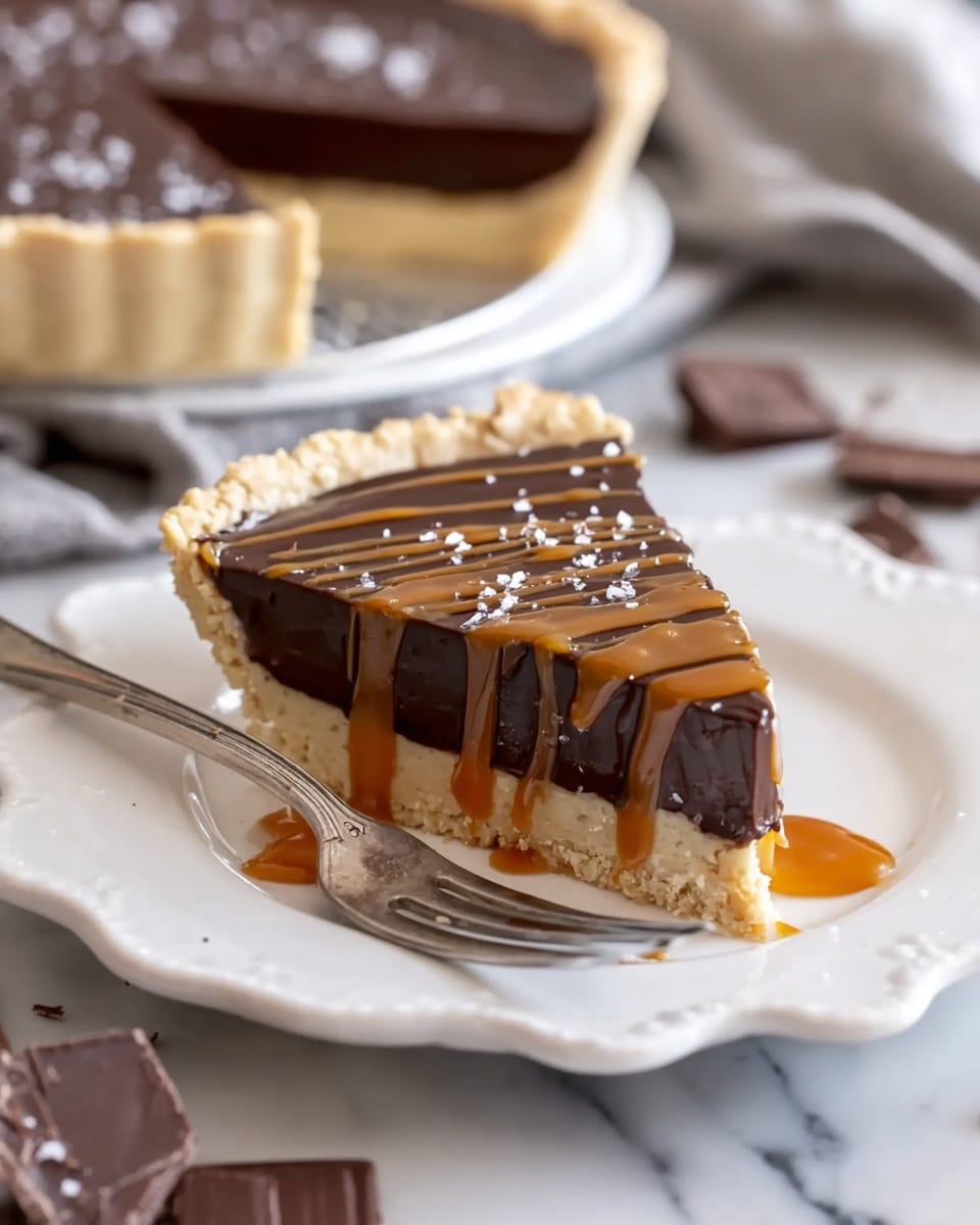 A slice of pie with three layers sits on a white plate with scalloped edges: a crumbly light beige crust at the bottom, a smooth dark chocolate filling in the middle, and a shiny caramel sauce drizzled on top in thin lines. There are small flakes of white salt sprinkled on the chocolate. A piece of the pie is on a fork in the front, showing the layers clearly with some caramel drizzled on it too. In the blurry background, the rest of the pie and some chocolate shavings can be seen on a white marbled surface. Photo taken with an iphone --ar 4:5 --v 7
