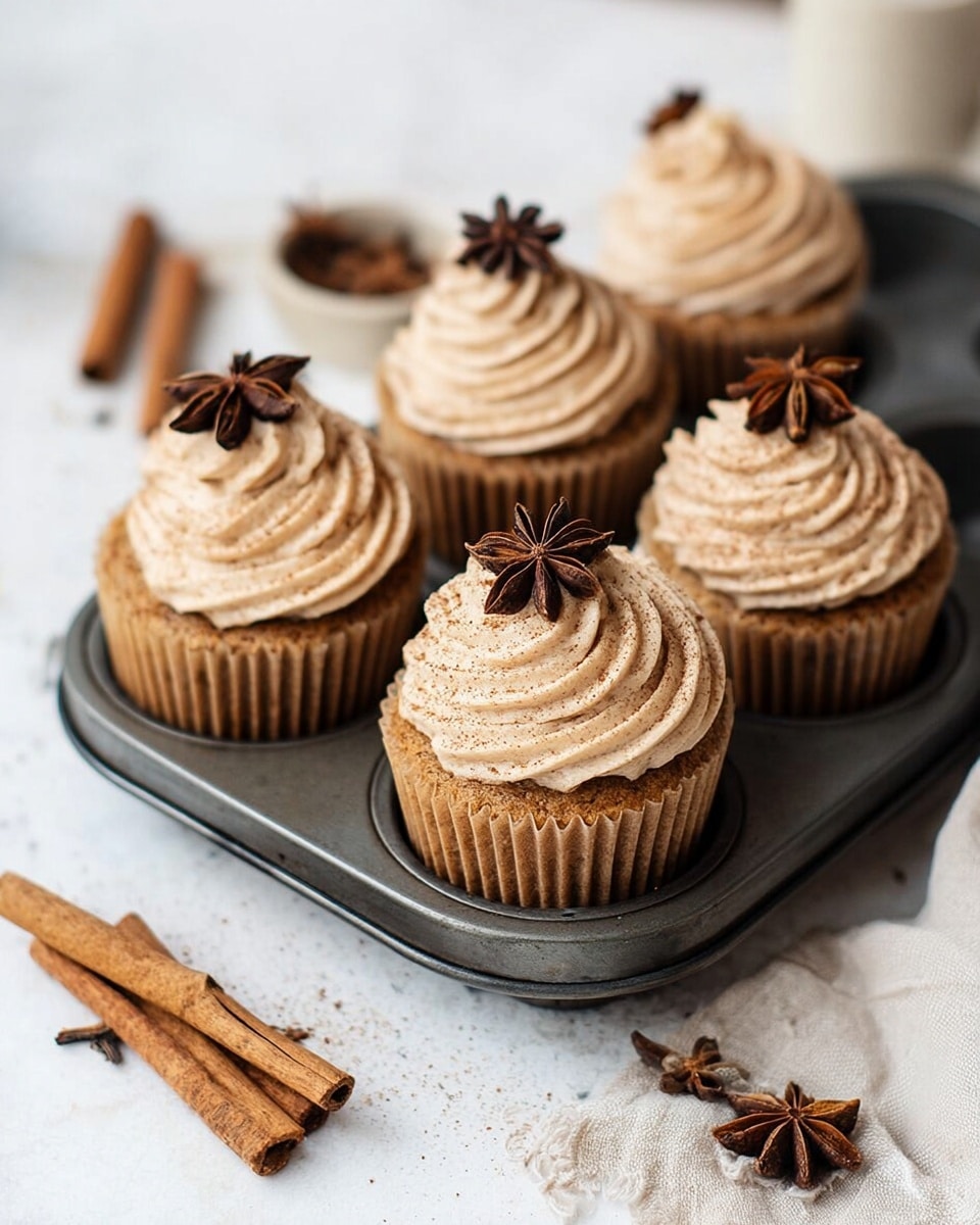 The image shows five cupcakes arranged in a metal cupcake tray, each topped with a swirl of light brown frosting that has a smooth, creamy texture. At the peak of each frosting swirl sits a single dark brown star anise, adding a decorative touch. The cupcakes have a golden brown base with a slightly rough texture, visible through a brown ridged paper cupcake liner. The tray rests on a crumpled beige cloth, with a cinnamon stick and cloves scattered in the foreground on a white marbled surface. An open book is blurred in the background, giving a cozy, warm atmosphere. photo taken with an iphone --ar 4:5 --v 7