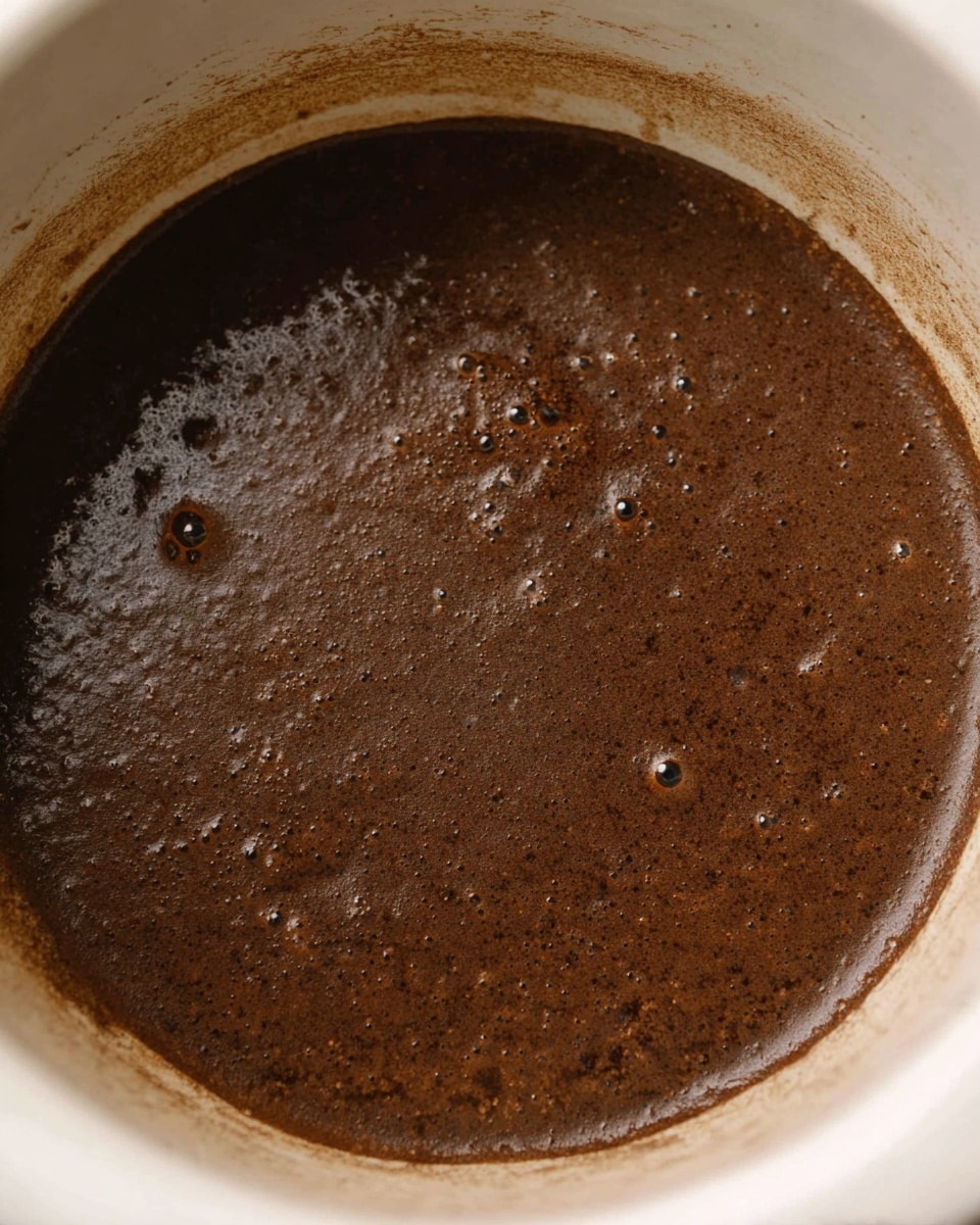 The image shows a close-up view of a single layer of thick, dark brown liquid with a slightly rough texture and small bubbles scattered on the surface, contained inside a white round pot with visible residue on the inner rim. The liquid has a matte finish with some areas reflecting light, creating a subtle shine, and it fills the pot almost to the edges. The background is not visible as the focus is entirely on the pot's interior and the rich brown liquid inside. photo taken with an iphone --ar 4:5 --v 7