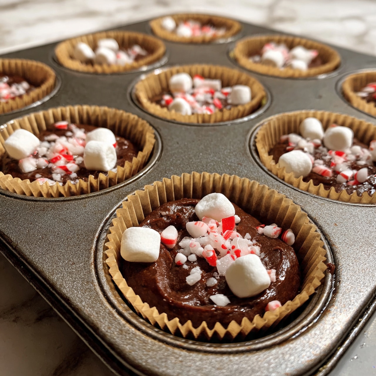 This image shows close-up of a dark grey muffin tin holding white paper cupcake liners filled with three layers, starting with a smooth, shiny dark brown chocolate batter base. The next layer is a scattering of small, soft white mini marshmallows sitting on top of the batter. In some cups further back, there is a mix of crushed red and white peppermint candy pieces layered over the dark chocolate batter instead of marshmallows. The muffin tin sits on a white marbled surface. Photo taken with an iphone --ar 4:5 --v 7