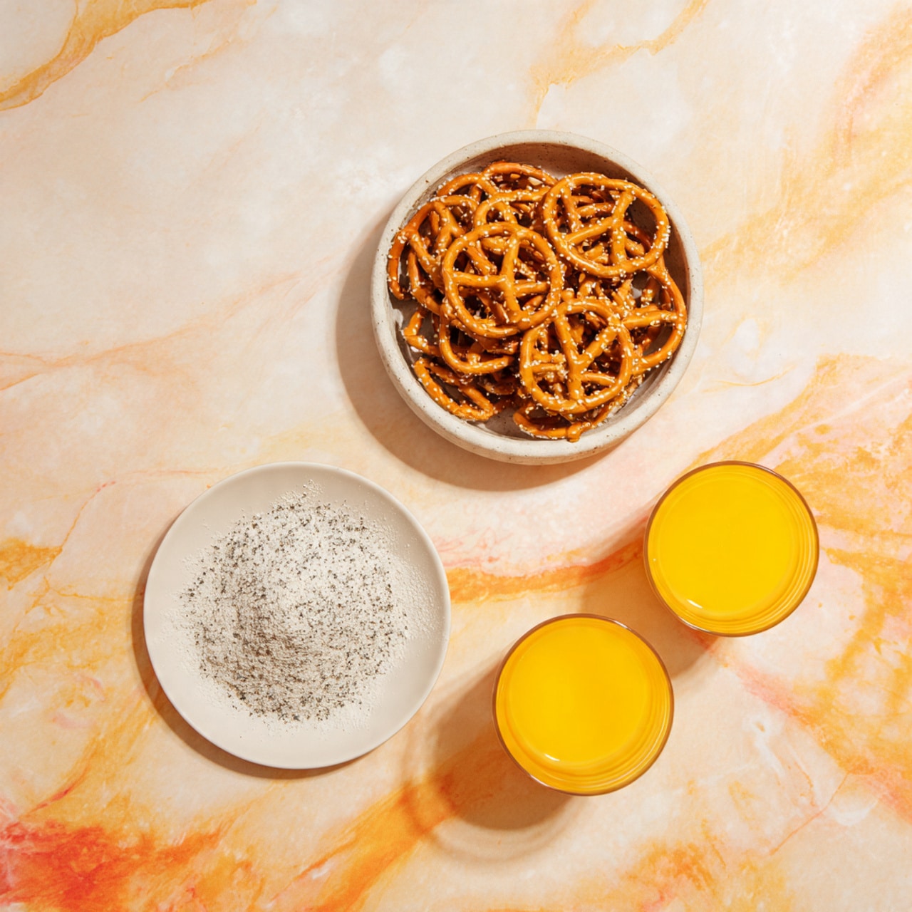 A round white bowl filled with many small golden brown pretzels stacked up to the top. Below the bowl, there is a small white plate containing a pile of fine white powder mixed with tiny black specks. On each side of the small plate, there are two clear glass cups filled with a bright yellow liquid. All items rest on a white marbled surface with soft orange and yellow tones around. Photo taken with an iphone --ar 4:5 --v 7