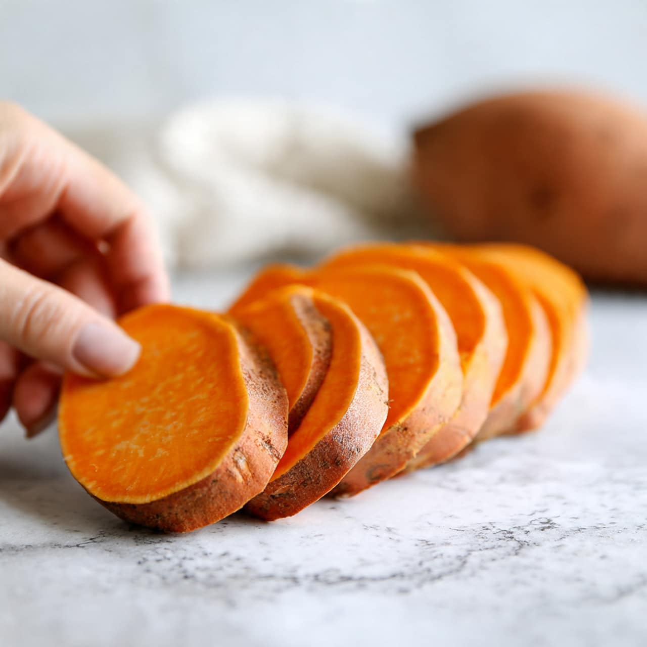 The image shows a close-up of several slices of sweet potato with a smooth, bright orange inside and a darker brown skin edge. They are placed in a slightly curved line on a white marbled surface. In the background, there is a whole sweet potato with its brown skin still on, softly out of focus. A blurry woman's hand is seen near the whole sweet potato, suggesting someone is about to take or has just placed it down. The scene has a bright, natural light. photo taken with an iphone --ar 4:5 --v 7