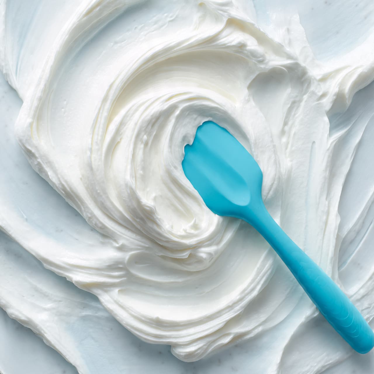 The image shows a close-up of smooth and fluffy white cream with soft peaks and swirls. A light blue silicone spatula is stuck into the cream, resting on its thick texture. The cream fills most of the view, and the background is a white marbled texture. The cream looks rich and well-whipped, with a soft, airy appearance. photo taken with an iphone --ar 4:5 --v 7