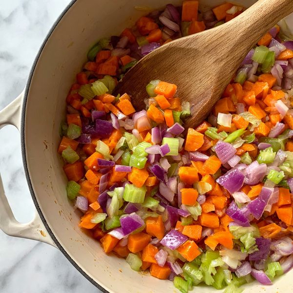 A close-up of a white pot with diced colorful vegetables cooking inside, including orange carrots, purple onions, and light green celery pieces. A wooden spoon stirs the mixture, showing the mix's moist and soft texture. The colors of the vegetables are bright and fresh, and there is some steam rising from the pot. The background shows a white marbled texture. Photo taken with an iphone --ar 4:5 --v 7