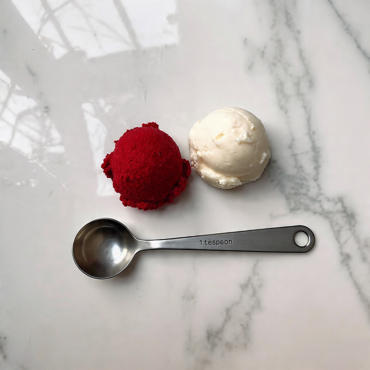 The image shows two small scoops of sorbet placed side by side on a white marbled surface. The scoop on the left is bright red with a smooth and slightly soft texture, while the scoop on the right is light cream-yellow with a slightly grainy texture. Below the scoops is a shiny metal measuring spoon marked