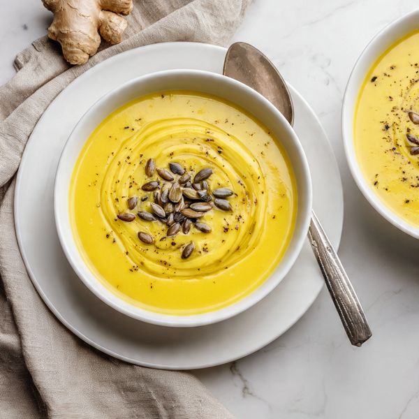 The image shows two white bowls filled with smooth, bright yellow soup, each placed on matching white plates. The main bowl in the center has a swirl of olive oil and a small pile of sunflower seeds with a sprinkle of black pepper on top. A silver spoon rests on the plate beneath the main bowl. There is a beige linen cloth folded near the bottom left, and a piece of fresh ginger is visible in the top left corner on a white marbled surface. The second bowl is partially visible in the lower right corner, also with black pepper on the soup. photo taken with an iphone --ar 4:5 --v 7