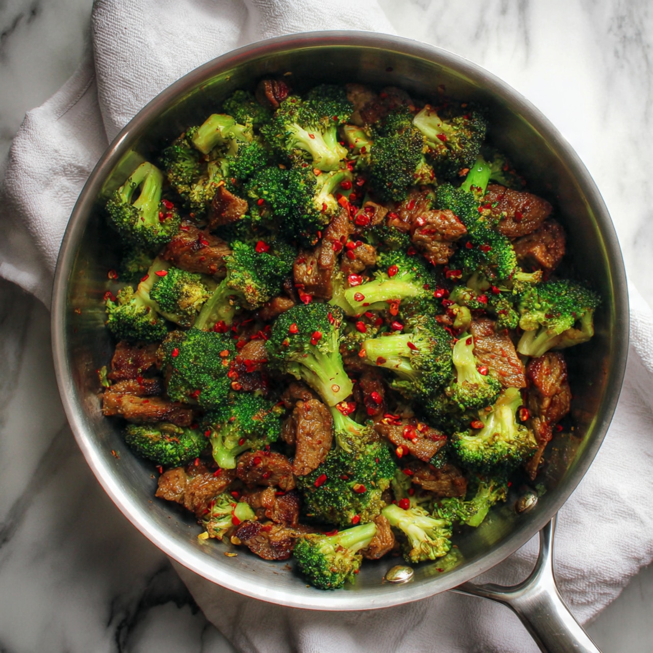 This image shows a stainless steel pan filled with a mix of cooked ground meat and chopped broccoli. The dish has many small pieces with a mix of green broccoli florets and light brown meat bits, evenly combined with visible red chili flakes scattered throughout. The pan sits on a light brown cloth over a white marbled surface. The colors mainly show green from the broccoli and light brown from the meat. photo taken with an iphone --ar 4:5 --v 7