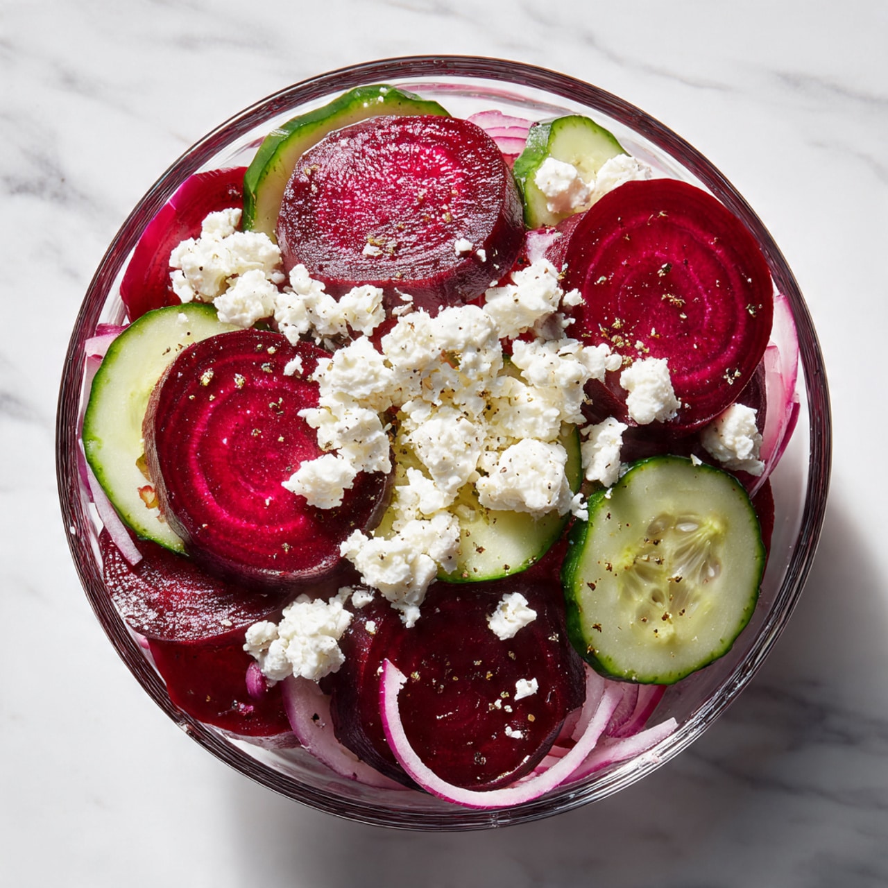 A clear glass bowl holds a layered mix of fresh salad ingredients on a white marbled surface. The bottom layer has thin slices of purple-red onion with a slightly translucent texture, placed to one side. Next to the onion, there are light green cucumber slices with darker green edges, stacked neatly in a row. On the other side, deep red, glossy beet slices fill the bowl. At the center top, small cubes of white feta cheese with crumbly texture are piled over the vegetables. Photo taken with an iphone --ar 4:5 --v 7