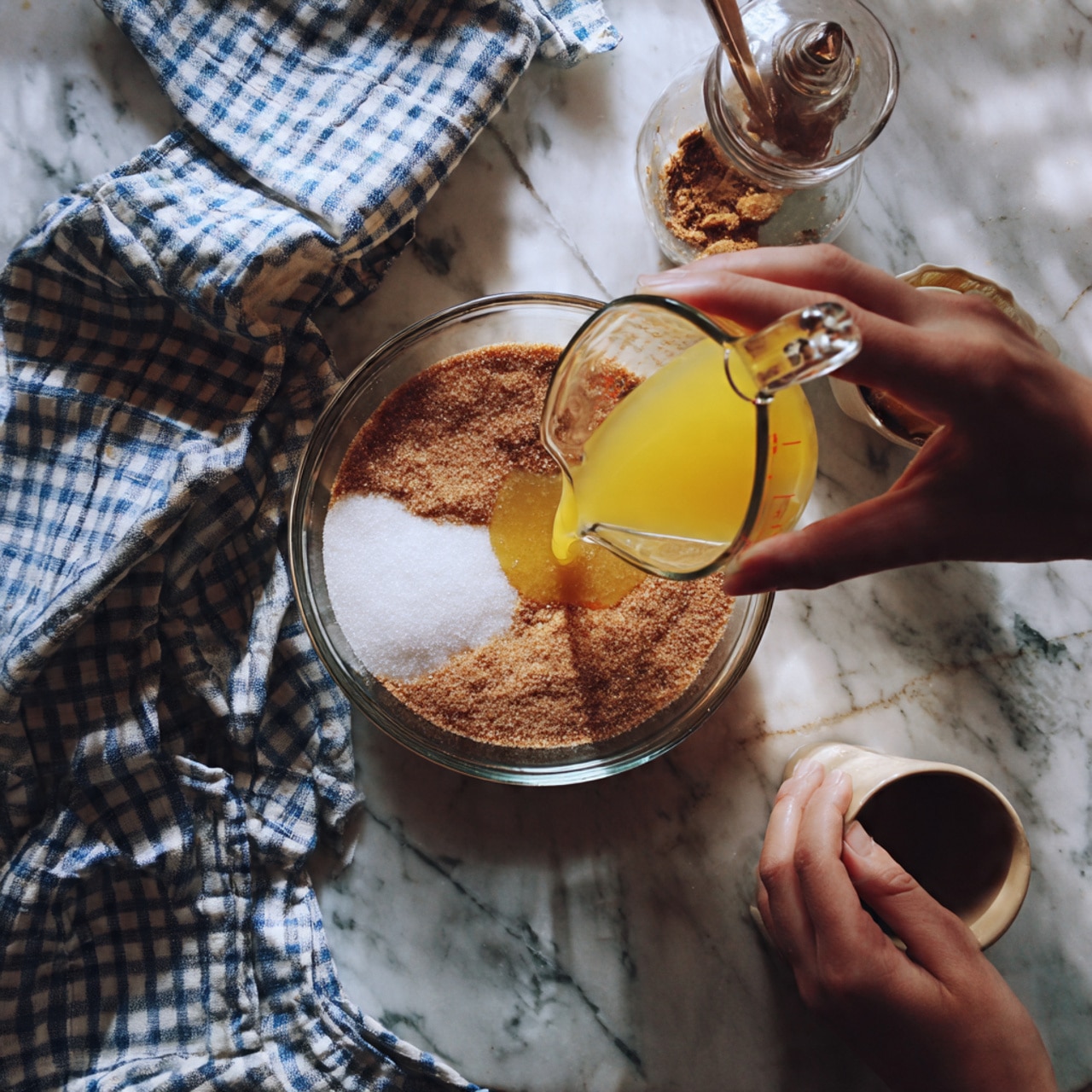 A clear glass bowl sits on a white marbled surface with a layer of white granulated sugar on the right side, a mound of brown sugar on the left, and a small pool of melted butter in the center. A woman's hand is pouring a stream of bright yellow liquid, likely melted butter or oil, from a clear glass measuring cup into the bowl. A small glass cup with a light brown liquid sits in the upper right corner, and a white cup filled with orange liquid rests on the lower left. A red and white checkered cloth is partially visible in the upper left background. Photo taken with an iphone --ar 4:5 --v 7