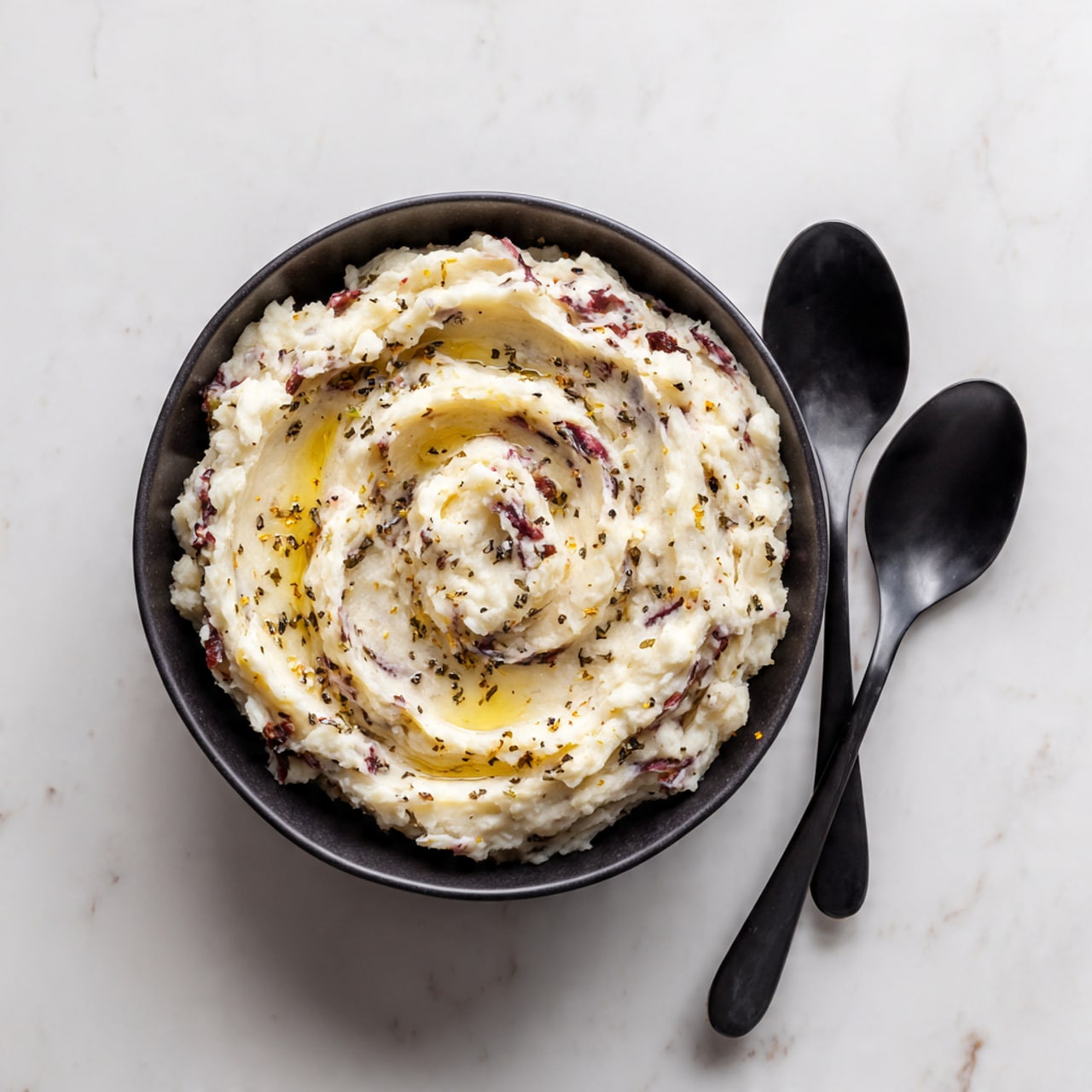 A black round bowl filled with creamy mashed potatoes that have bits of red potato skin mixed throughout. The mash is swirled with a melting pat of butter in the center, which is slightly shiny and smooth. Tiny black pepper specks are scattered on the surface. On the right side of the bowl, two black spoons rest, with a small dollop of mashed potatoes on one of them. The bowl sits on a white marbled surface. photo taken with an iphone --ar 4:5 --v 7