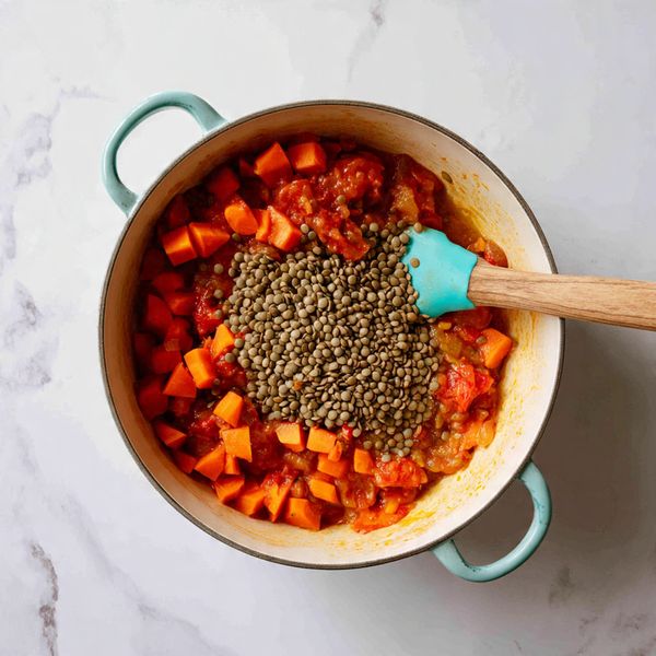 The image shows a white pot filled with a vibrant mix of ingredients, mostly orange and red in color from diced carrots and tomato pieces in a thick sauce base. On top, there is a large pile of dry green lentils covering the center. A turquoise spatula is resting inside the pot on the right side, partially submerged in the mix. A wooden spoon is stirring from the left side. The pot is placed on a white marbled surface. photo taken with an iphone --ar 4:5 --v 7