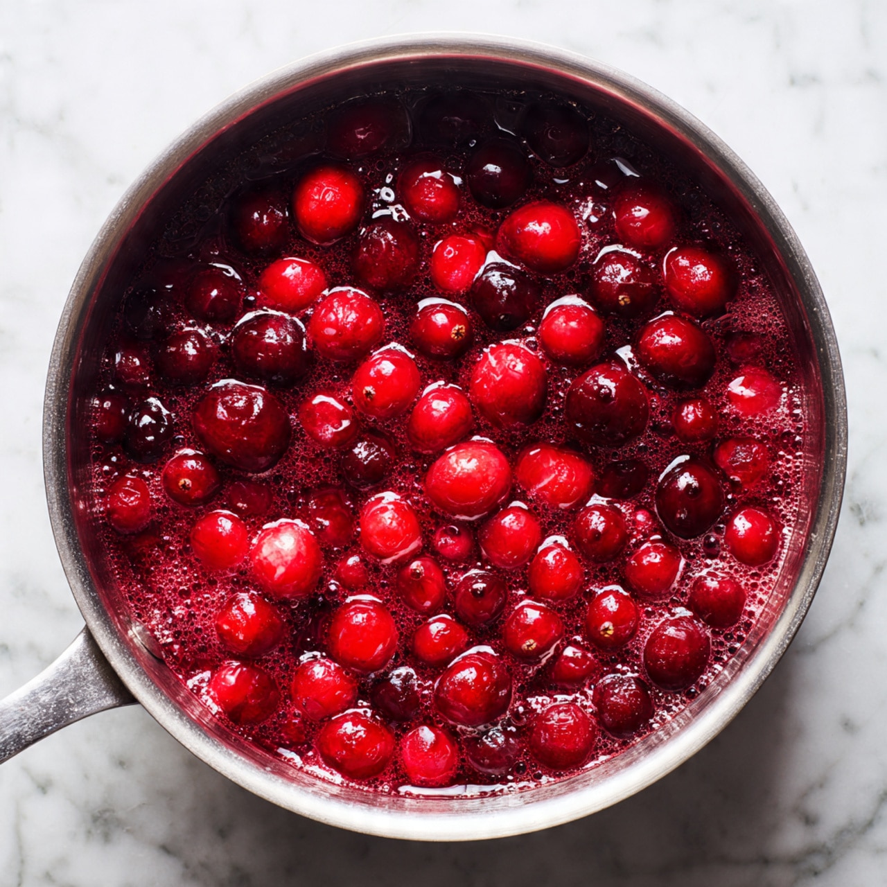 A metal pot filled with bright red cranberries cooking in a thick, syrupy liquid that bubbles and steams, giving a shiny and wet texture to the berries. The cranberries are whole and some are breaking open, showing a mix of deep red and lighter red shades, resting against the silver edges of the pot on a stove, with a faint blurred background. The scene sits on a white marbled surface. photo taken with an iphone --ar 4:5 --v 7