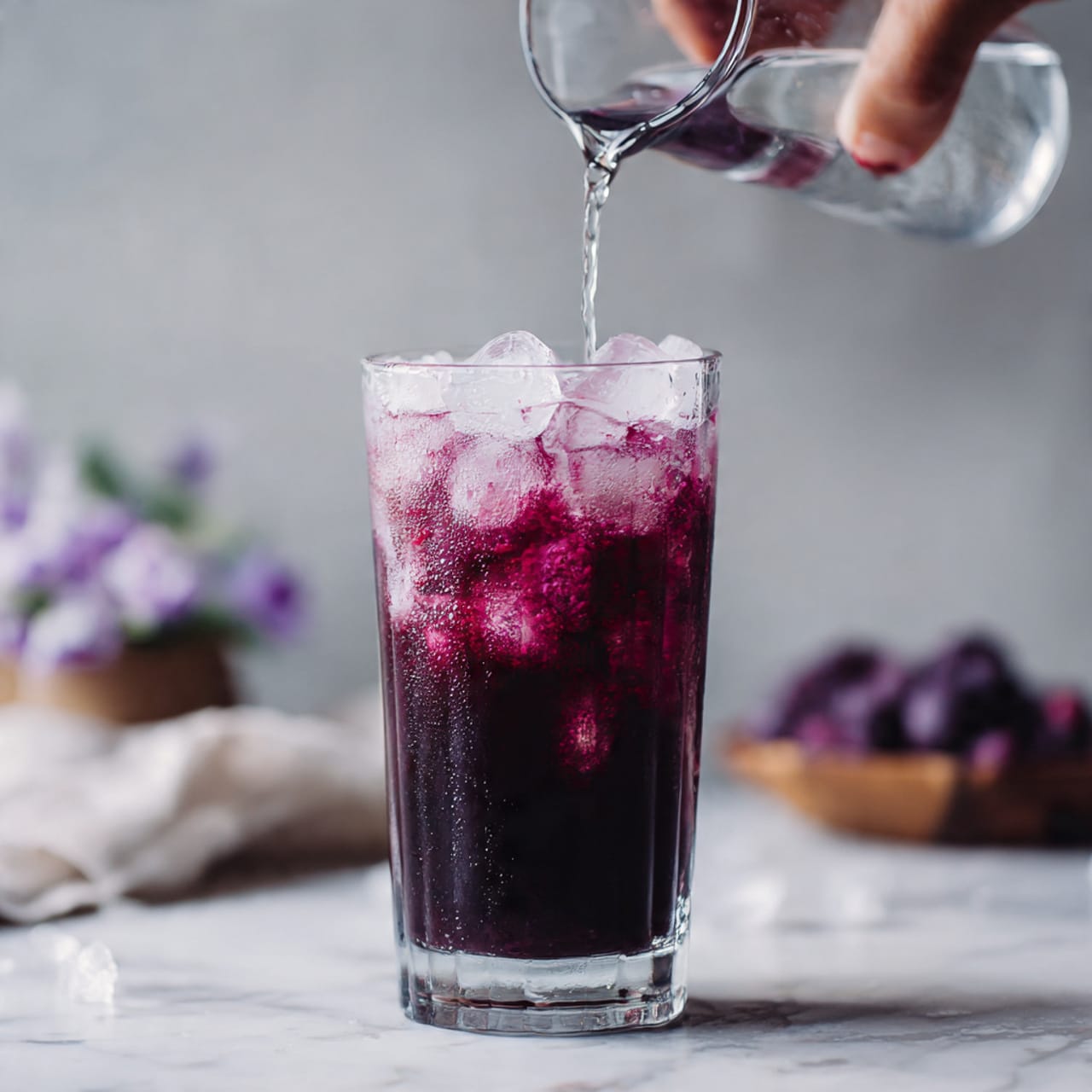 A tall clear glass filled with dark purple iced drink fading to a lighter purple near the top, with several ice cubes inside. A woman's hand is pouring clear liquid from a glass container into the drink. The background and surface have a white marbled texture. Photo taken with an iphone --ar 4:5 --v 7