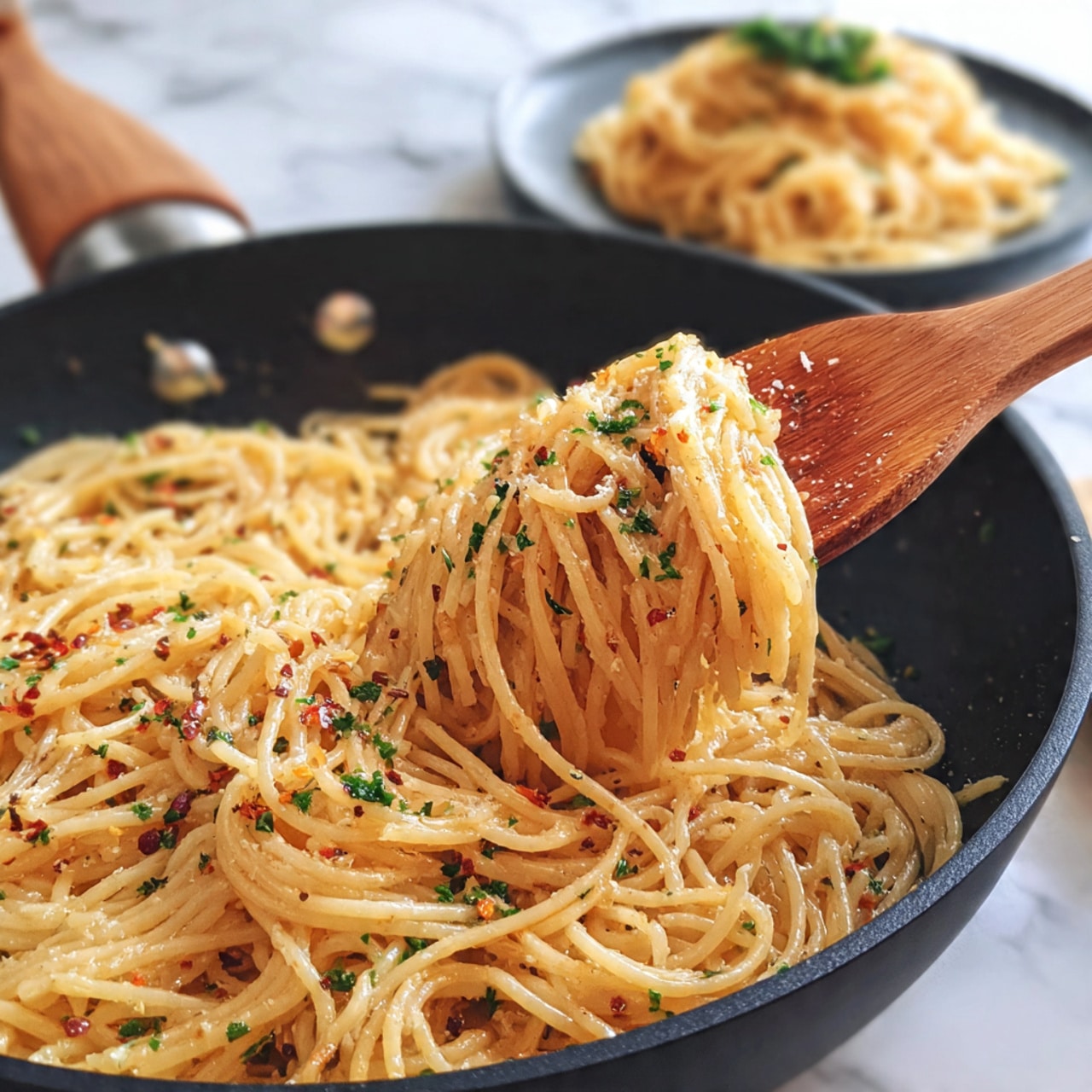 The image shows one layer of cooked spaghetti pasta mixed with small green herb pieces and red chili flakes, all inside a dark pan. Above the pan, a wooden spoon lifts a portion of the pasta, showing many long, thin, yellow noodles hanging down with bits of green herbs and red chili flakes scattered throughout. In the background, there is a blurred white plate with more spaghetti. The setting is on a white marbled surface. Photo taken with an iphone --ar 4:5 --v 7