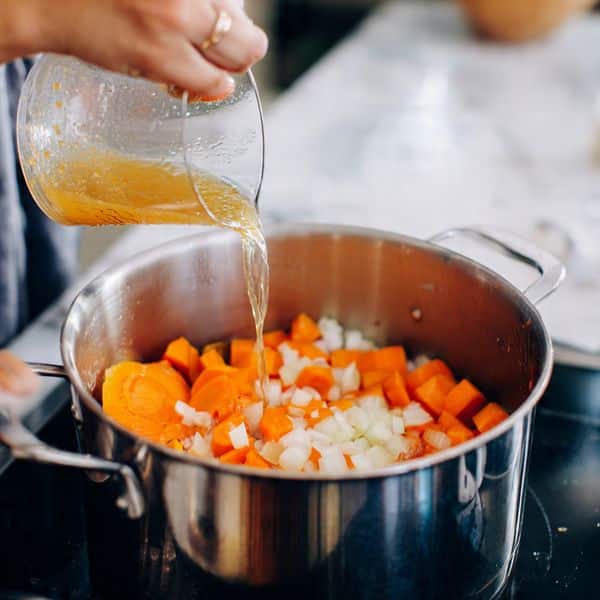 A close-up image showing a woman's hand pouring light golden broth from a clear measuring cup into a large stainless steel pot filled with thick orange carrot chunks and small white onion pieces. The pot sits on a black stovetop, and the background surface is white marbled texture. The colors show the vibrant contrast between the bright carrots, the clear broth, and the shiny metal pot. Photo taken with an iphone --ar 4:5 --v 7