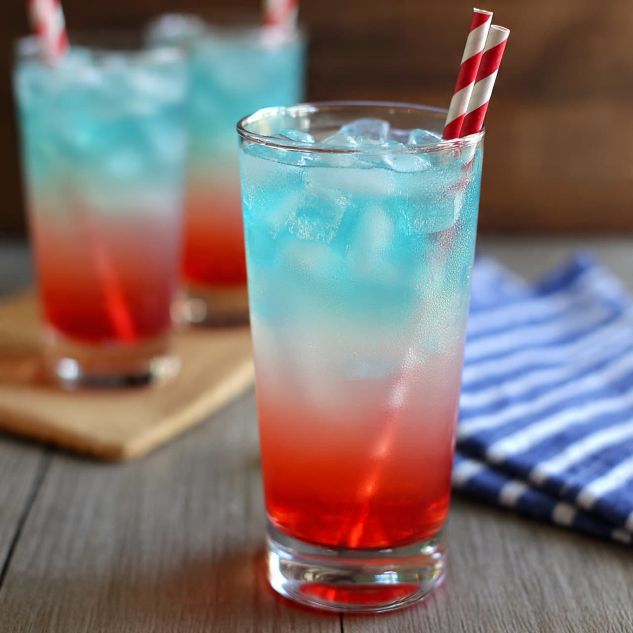 A tall clear glass filled with three layers of colorful drink: the bottom layer is bright red, the middle layer is a soft pink blending up, and the top layer is light blue with many large ice cubes. Two red and white striped straws stand in the glass. In the background, two similar glasses are slightly out of focus on a light brown wooden surface with a blue and white striped cloth nearby. Photo taken with an iphone --ar 4:5 --v 7