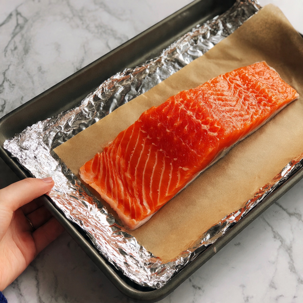A bright orange salmon fillet with visible white lines runs diagonally from top left to bottom right. It rests on a white baking tray lined with white parchment paper and surrounded by crinkled silver aluminum foil edges. The tray is placed on a smooth white marbled surface with subtle grey veins. The salmon's texture is smooth and shiny, with a natural pattern of stripes. Photo taken with an iphone --ar 4:5 --v 7