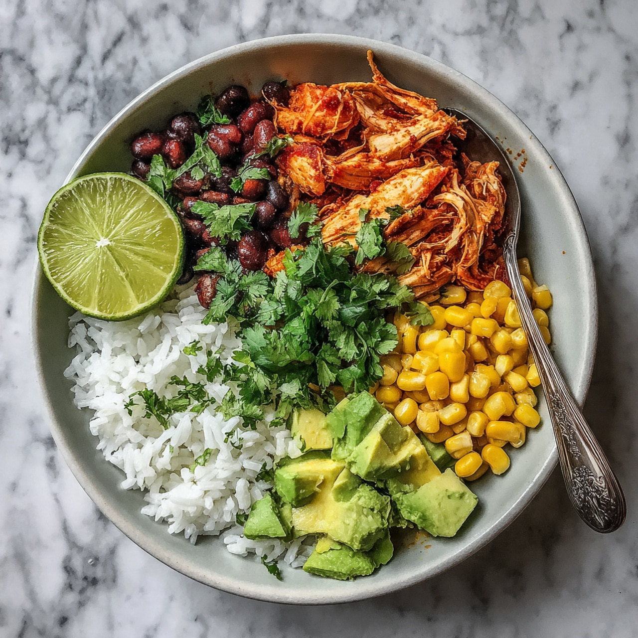 A white bowl sits on a white marbled surface, filled with layers of food. The bottom layer is fluffy white rice. On top, there is a central pile of shredded orange chicken mixed with small bits of red pepper, drizzled with a light green sauce, and sprinkled with fresh green cilantro leaves. Surrounding the chicken are four sections: diced green avocado on the top left, small yellow corn kernels on the right, and black beans on the left side. A wedge of bright green lime rests on the rice near the corn. A fork is placed inside the bowl on the right side. A piece of cilantro is on the marbled surface near the bowl, and a striped cloth is blurred in the background. Photo taken with an iphone --ar 4:5 --v 7