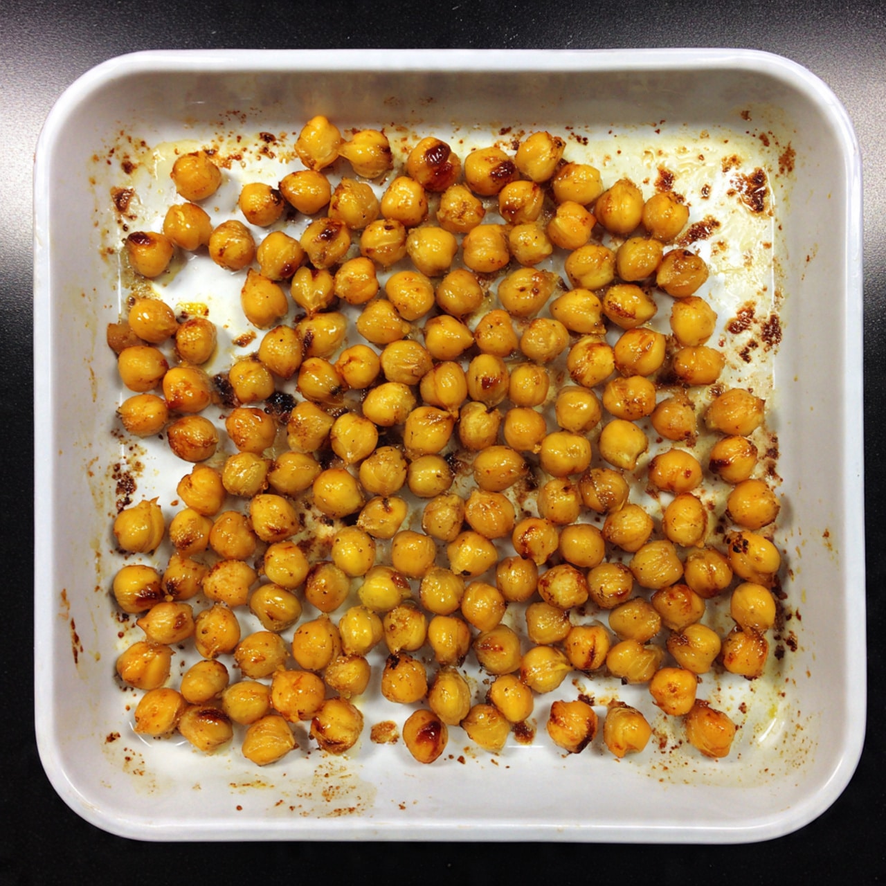 The image shows a shallow white baking tray filled with one even layer of small, round roasted chickpeas. They are golden brown with a slightly crispy texture, some chickpeas showing darker brown spots. The tray has some light browned bits and oil stains scattered around, indicating they have been roasted. The background is a white marbled surface. photo taken with an iphone --ar 4:5 --v 7