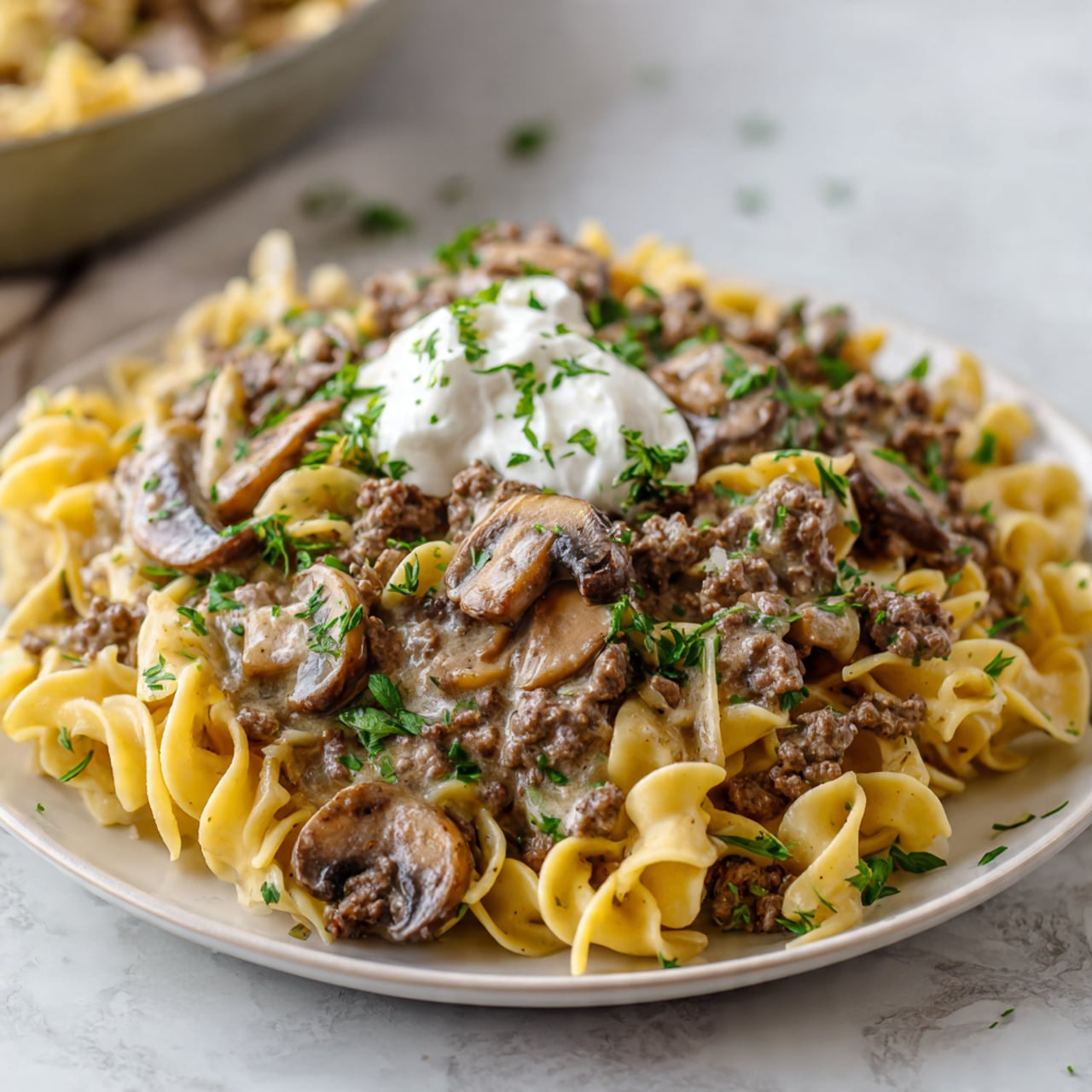 The image shows a white plate filled with three layers of food. The bottom layer is a pile of twisted, yellowish egg noodles with a slightly rough texture. On top of this is a thick, creamy brown sauce with pieces of cooked ground meat and sliced mushrooms, some of which are round and brown with a soft look. The dish is garnished with small green herb pieces scattered on top, and a small dollop of white cream sits in the middle. The plate is set on a white marbled surface, and the background is softly blurred with a large round pan of the same food visible. Photo taken with an iphone --ar 4:5 --v 7