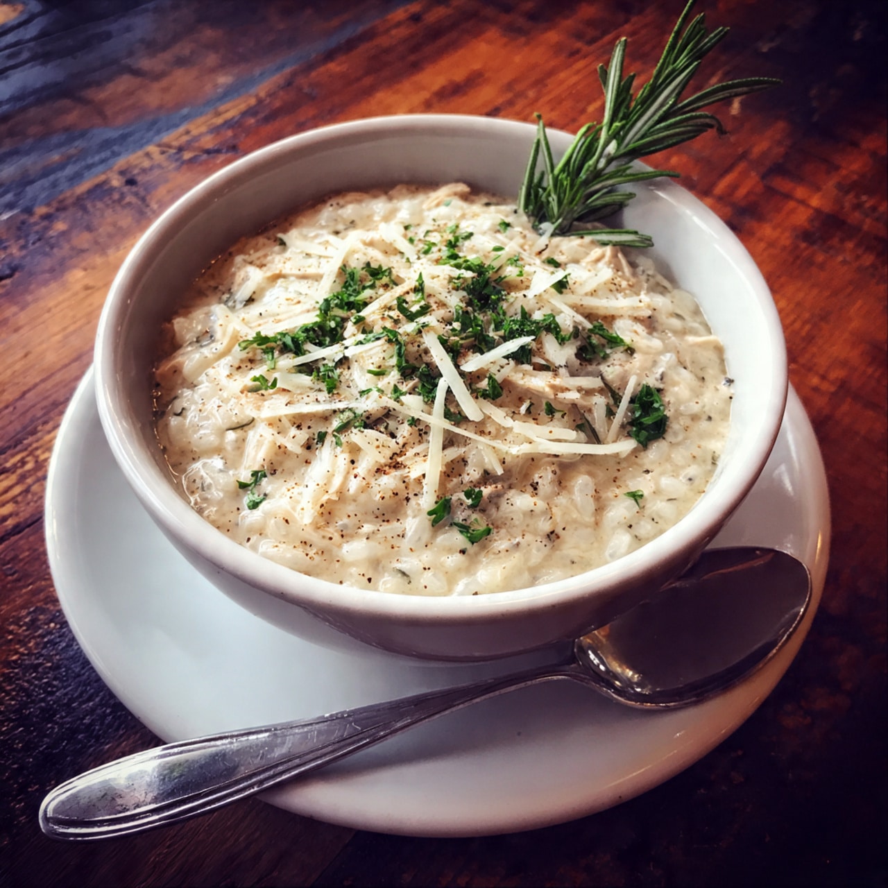 A close-up view of a bowl filled with creamy risotto featuring visible pieces of orange carrots, pale green celery, and tender small light brown chicken chunks. The risotto has a thick, textured creamy white base with a slight shine. The top is sprinkled with small green parsley leaves and thin off-white shavings of parmesan cheese. A sprig of fresh green rosemary is placed upright on one side of the bowl. The bowl is white and sits on a white marbled surface, with a white bowl partially visible in the background. photo taken with an iphone --ar 4:5 --v 7