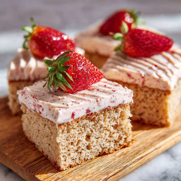 A square piece of light brown cake with small red specks is on a white plate with subtle textured lines. The cake has two layers: a thick, moist base and a creamy top layer with a pinkish swirl of strawberry sauce. On top, there is a whole bright red strawberry with green leaves. In the blurred background, more pieces of the same cake can be seen on a white marbled surface. A silver fork rests on the edge of the plate, and small white flowers are in the foreground. photo taken with an iphone --ar 4:5 --v 7