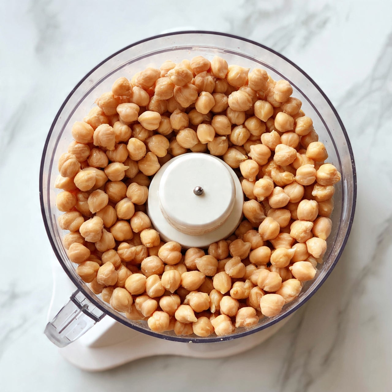 This image shows a clear food processor bowl filled with a single layer of light brown chickpeas. The chickpeas are round and smooth, closely packed around the white central blade inside the processor. The background surface has a white marbled texture, giving a clean and bright look. The food processor is seen from above, with the chickpeas evenly spread in a circle inside the bowl. Photo taken with an iphone --ar 4:5 --v 7