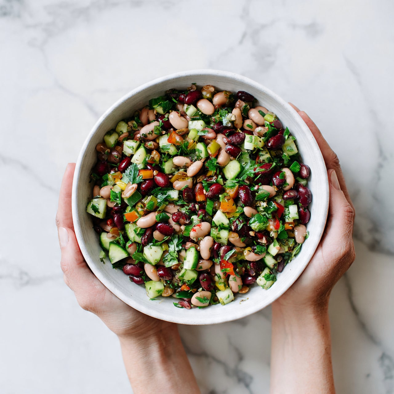 A white bowl held by two woman's hands is filled with a colorful bean salad. The salad has three layers mainly made up of different beans in light brown, dark brown, and beige colors. Mixed with the beans are bright green cucumber pieces and chopped fresh herbs that add a leafy texture. The salad looks fresh and vibrant, with a mix of round and small dice shapes, all sitting on a white marbled surface. photo taken with an iphone --ar 4:5 --v 7