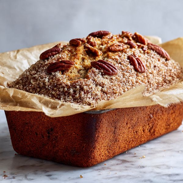 A rustic loaf of bread or cake in a parchment paper-lined rectangular pan, topped with a rough layer of crunchy crumb coating mixed with sugar and cinnamon, and scattered whole and halved pecan nuts partly embedded in the crumb topping. The loaf is golden brown with a cracked, uneven surface showing a soft interior under the crunchy coating. The parchment paper is slightly crinkled around the edges, and the background is a white marbled texture. Photo taken with an iphone --ar 4:5 --v 7