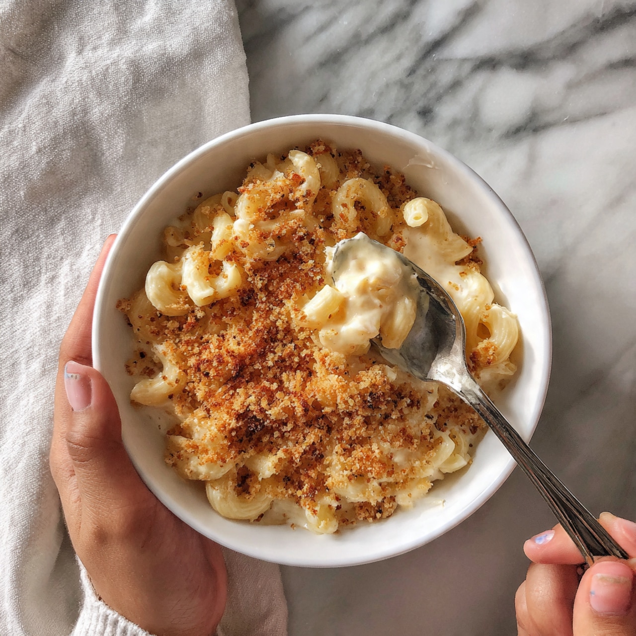 The dish is a creamy pasta served in a white bowl, filled with small elbow macaroni noodles covered in a thick white cheese sauce. On top, there is a crispy golden-brown layer of breadcrumbs with hints of green herbs sprinkled around for color. A woman’s hand is holding a silver spoon scooping into the pasta, showing the creamy texture beneath the crunchy topping. The background features a white marbled surface. photo taken with an iphone --ar 4:5 --v 7
