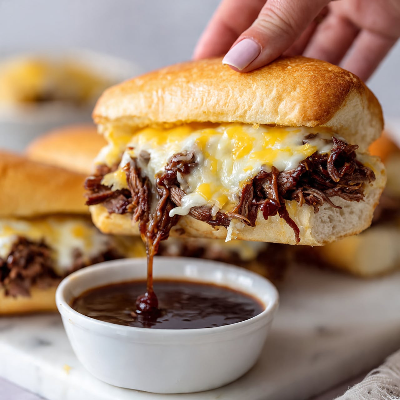 A close-up image shows a sandwich held by a woman's hand, with a white crusty sandwich roll on top. Inside the sandwich, there is a thick layer of shredded dark brown meat mixed with melted light yellow cheese and some creamy melted cheese over the meat. The sandwich is held above a small white bowl filled with dark brown dipping sauce, with the sauce dripping slightly from the sandwich. The background is a white marbled surface with some blurred sandwiches in the back. Photo taken with an iphone --ar 4:5 --v 7