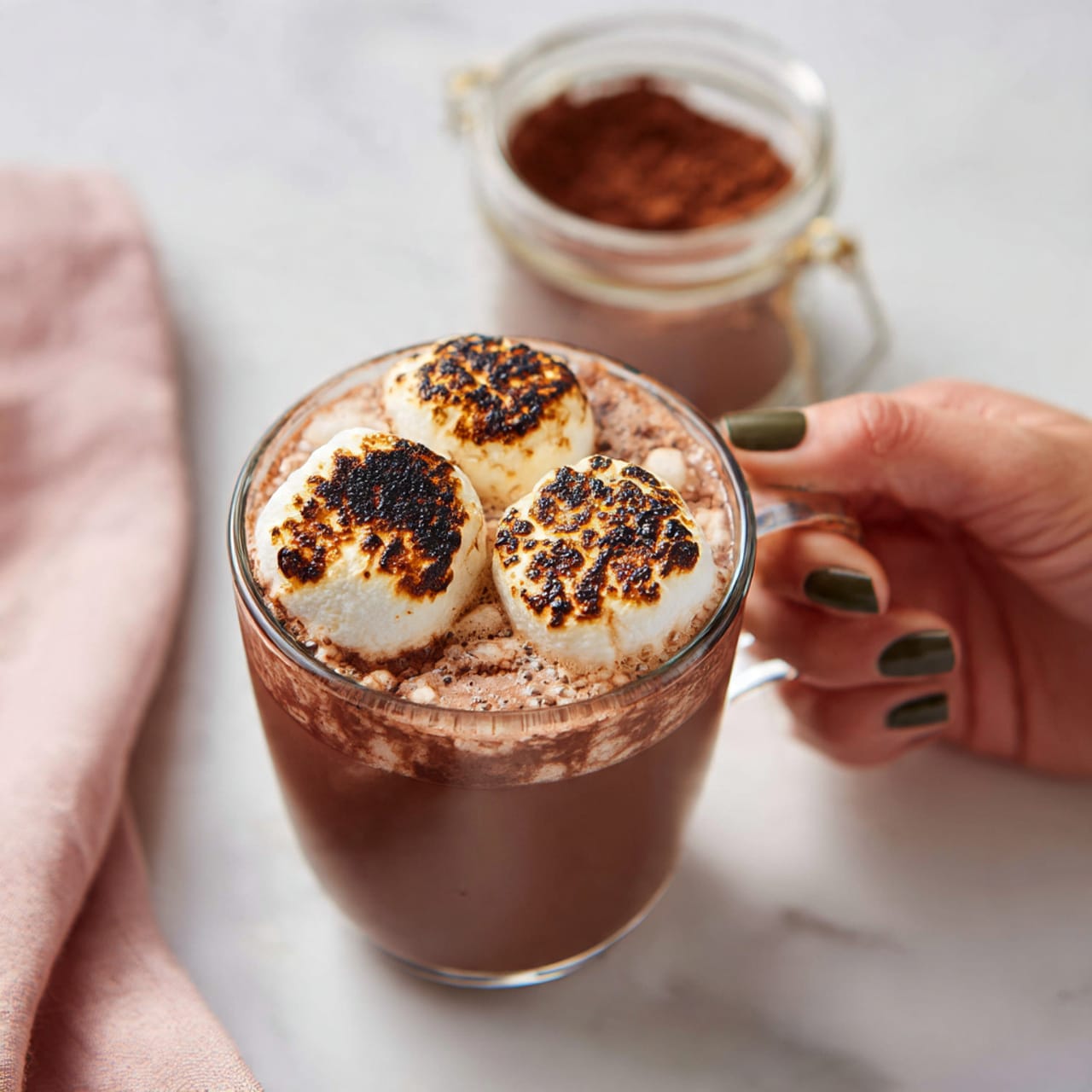 A clear glass cup holds rich, brown hot chocolate, topped with three large toasted marshmallows that have golden and dark brown spots from toasting. The cup is held by a woman's hand with dark green nails. The cup sits on a white marbled surface, and next to it is a glass jar filled with cocoa powder. A soft pink cloth is partly visible on the left side of the image. The scene feels cozy and warm, focused on the drink and its toasted marshmallow topping. photo taken with an iphone --ar 4:5 --v 7
