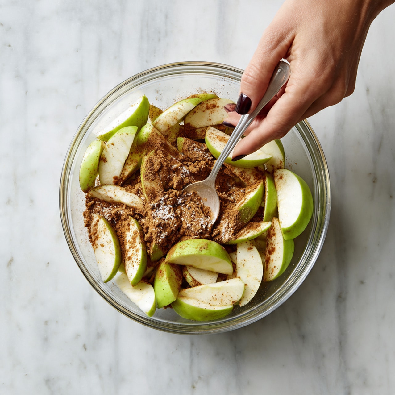 A clear glass bowl filled with many apple slices that are pale green and white in color, layered loosely and covered with scattered light brown cinnamon powder and white sugar granules. A woman's hand with dark-painted nails holds the edge of the bowl, while the other woman's hand stirs the contents with a spoon inside the bowl. The bowl sits on a white marbled surface. photo taken with an iphone --ar 4:5 --v 7