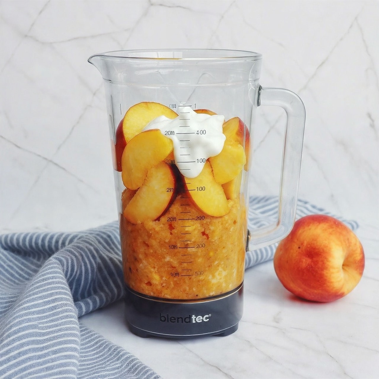 A tall clear blender jar sits on a white marbled surface with a soft blue and white striped cloth underneath. Inside the jar, there are three visible layers: at the bottom is a thick yellow-orange fruit mixture, above that are several whole peach slices arranged loosely, and the top layer is a dollop of white yogurt slightly melting into the fruit. The blender jar has black measurement markings on the side and the word