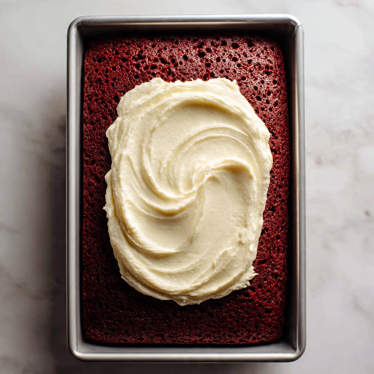 The image shows a rectangular baked red velvet cake in a silver metal baking tray, placed on a white marbled surface. The cake is deep red with small holes evenly spread on the top. A large dollop of thick, creamy white frosting sits in the center of the cake, ready to be spread. The texture of the cake looks moist and soft, contrasting with the smoothness of the frosting on top. photo taken with an iphone --ar 4:5 --v 7