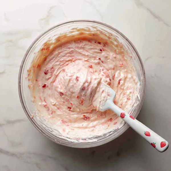 This image shows a glass bowl filled with thick, pale pink batter mixed with small red strawberry pieces, creating a textured and slightly chunky look. A white spatula with red strawberries printed on it is partially dipped into the batter, with some batter coated on the spatula's surface. The bowl is placed on a white marbled surface, and the texture of the batter looks creamy and well blended, ready to be used for baking photo taken with an iphone --ar 4:5 --v 7