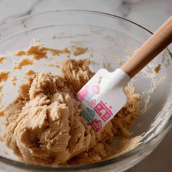A close-up view of a large clear bowl filled with light brown dough that looks soft and thick. The dough has a slightly grainy texture and is pushed upward by a white spatula with a wooden handle and pink and blue house designs on its silicone part, visible at the bottom right. The bowl sides have smears of dough stuck to them, showing the action of mixing. The background surface is a white marbled texture photo taken with an iphone --ar 4:5 --v 7