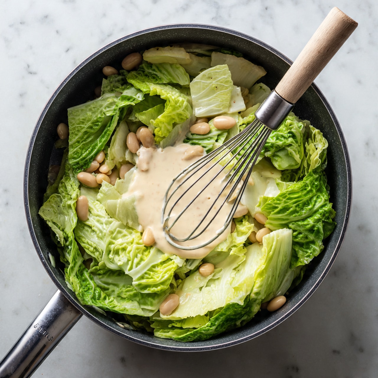 The image shows a black saucepan filled with a mix of fresh green cabbage leaves cut into large pieces scattered on top with light beige creamy sauce being whisked in the center by a silver whisk with a light wooden handle; small white beans are mixed under the cabbage leaves, and the inside of the pan has a dark non-stick texture. The saucepan is placed on a white marbled surface. photo taken with an iphone --ar 4:5 --v 7