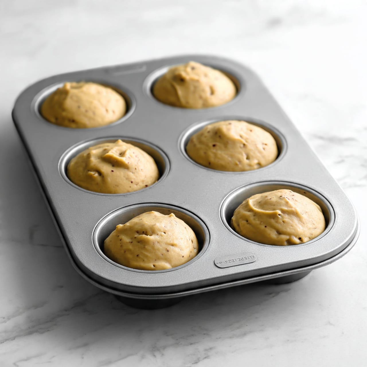 The image shows a gray metal muffin tray with six cavities, each cavity filled with a smooth, golden brown dough pressed into a shallow cup shape. The dough has a slightly textured surface with small dark specks, and each cup has a rounded bottom and slightly raised edges that fit snugly inside the tray wells. The tray is placed on a white marbled surface, and no other items are visible. photo taken with an iphone --ar 4:5 --v 7