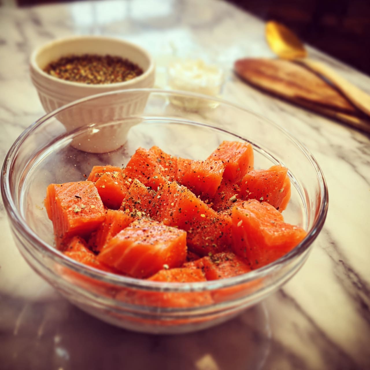 A clear glass bowl sits on a white marbled surface filled with about 18 bright orange salmon pieces arranged in a loose circle with some overlapping in the center. On top of the salmon pieces, there is a sprinkling of coarse salt and cracked black pepper creating white and black specks contrasting with the orange color. Above the bowl, slightly blurred but still visible, is a small white bowl containing white salt and black peppercorns arranged in a crescent shape. Photo taken with an iphone --ar 4:5 --v 7
